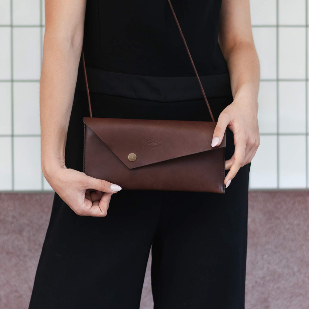 Woman holding a brown leather clutch bag with shoulder strap against a neutral background