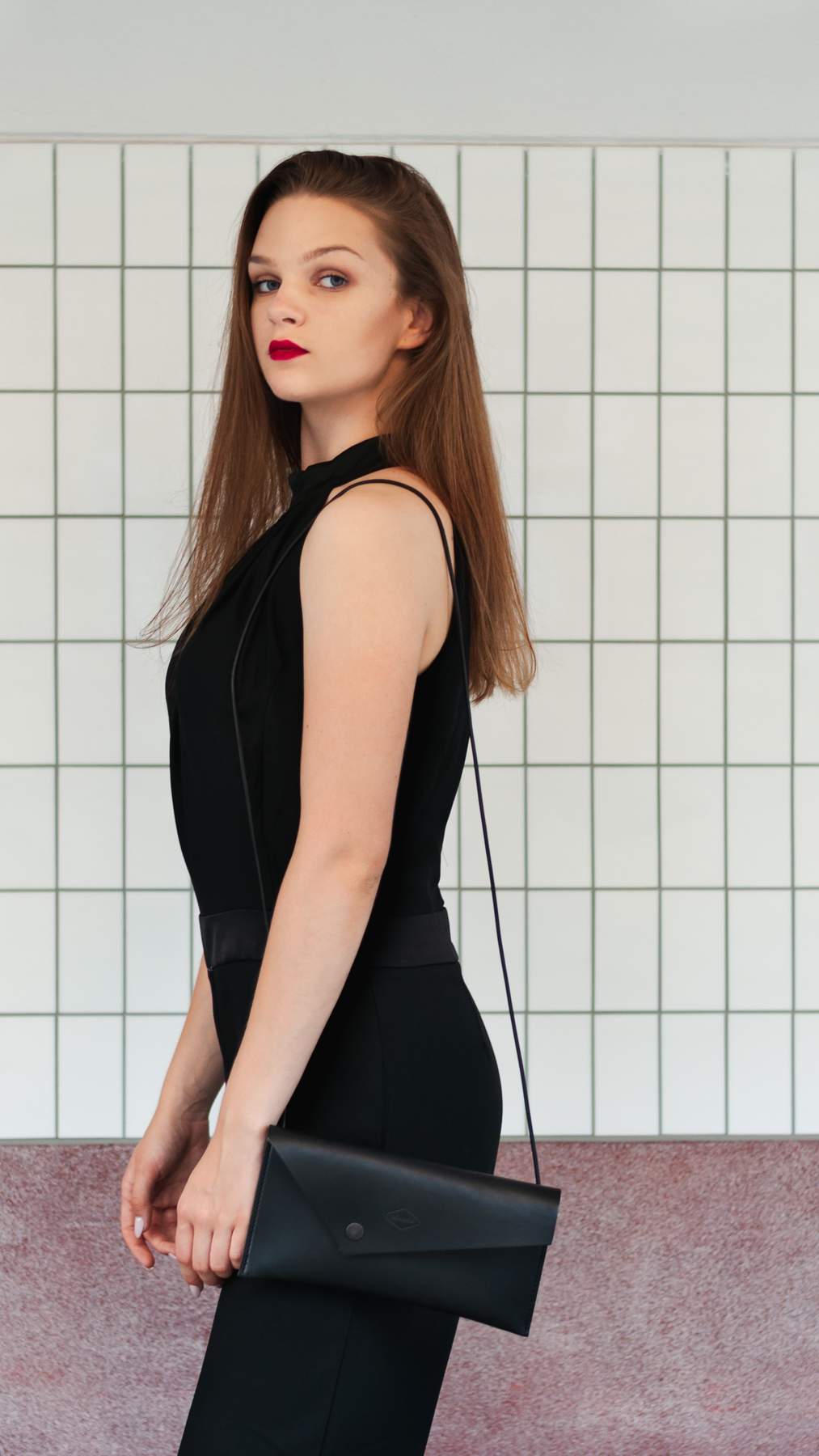 Woman in a black dress with a black clutch with shoulder strap against a tiled wall.