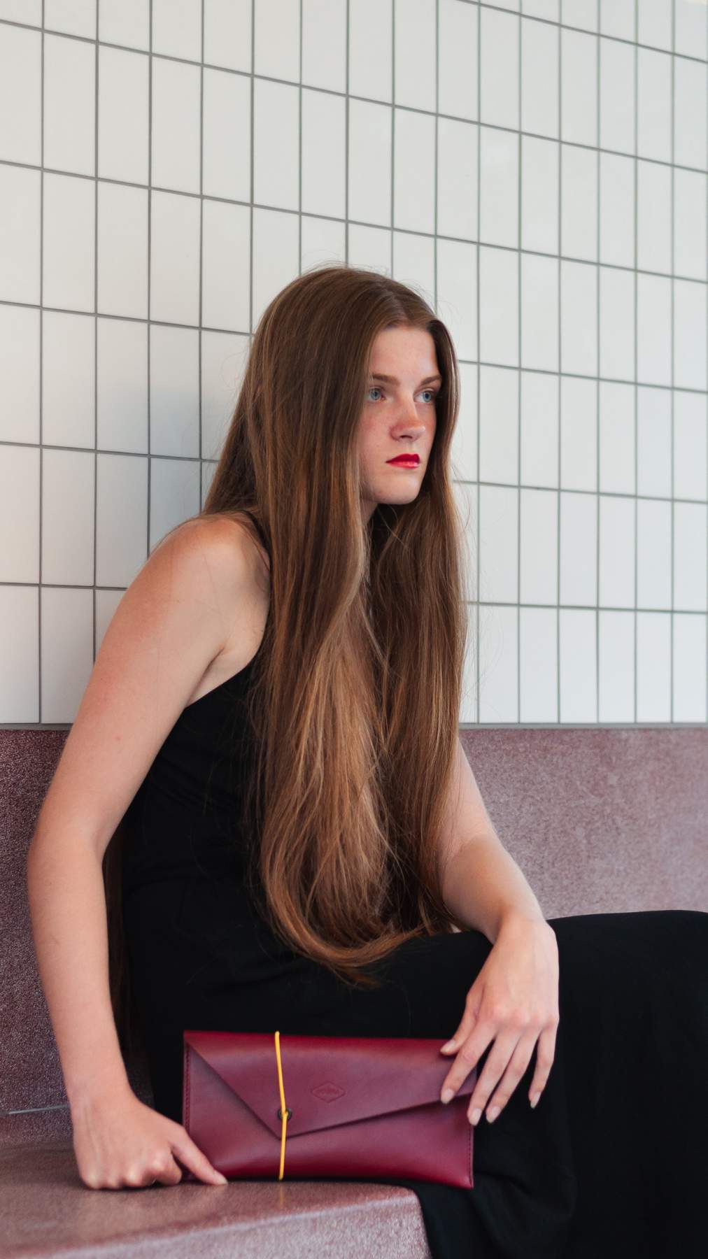 Woman holding a bordo clutch against a tiled wall.