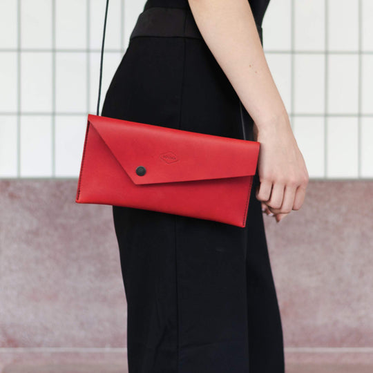 Red clutch bag with shoulder strap held by a person wearing black pants against a neutral background